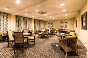 A hotel dining area with multiple tables and chairs, a patterned carpet, and framed pictures on the walls.