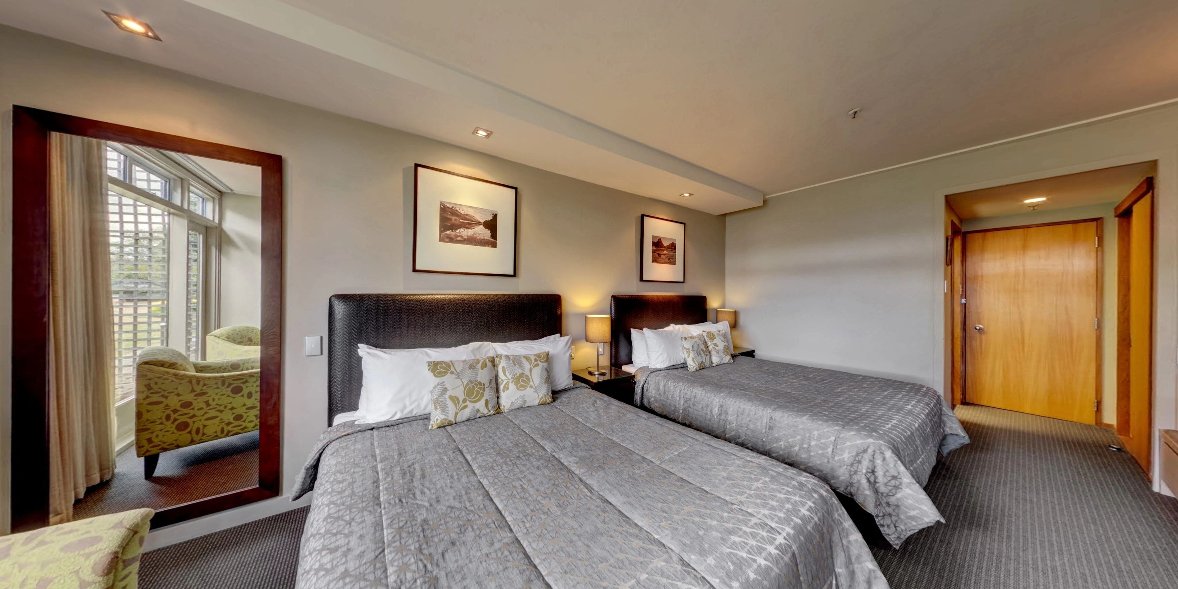 A hotel room with two beds, a large window with curtains, a chair, and framed pictures on the wall.
