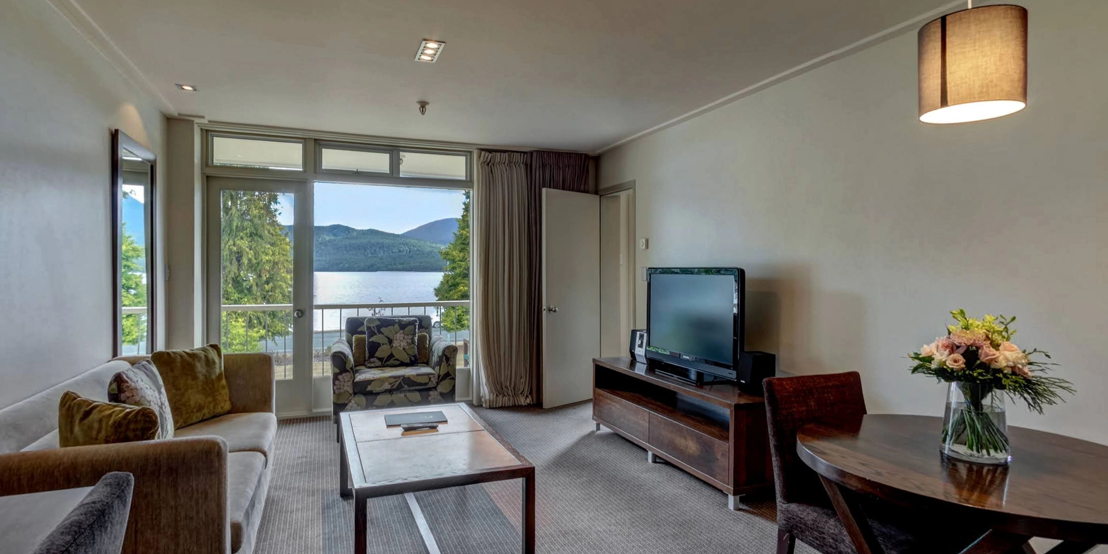 A hotel room with a sofa, a coffee table, a TV on a wooden stand, a dining table with chairs, a vase of flowers, and a balcony with a view of the lake and mountains.