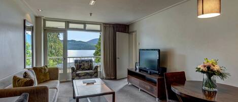 A hotel room with a sofa, a coffee table, a TV on a wooden stand, a dining table with chairs, a vase of flowers, and a balcony with a view of the lake and mountains.
