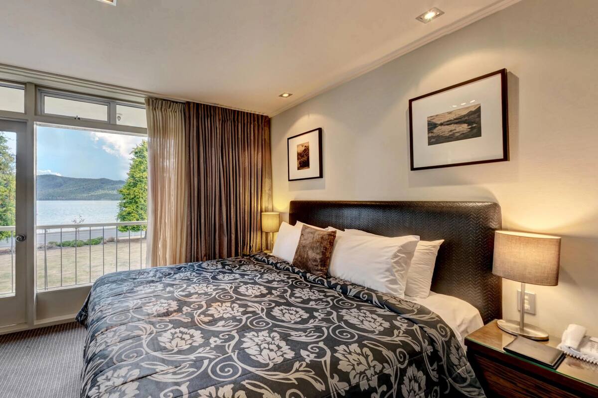 A hotel room with a large bed, a nightstand with a lamp, two framed pictures on the wall, and a balcony with a view of the mountains.