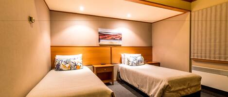 A hotel room with two single beds, wooden headboards, a small bedside table with a lamp, a painting on the wall, and a window with blinds.