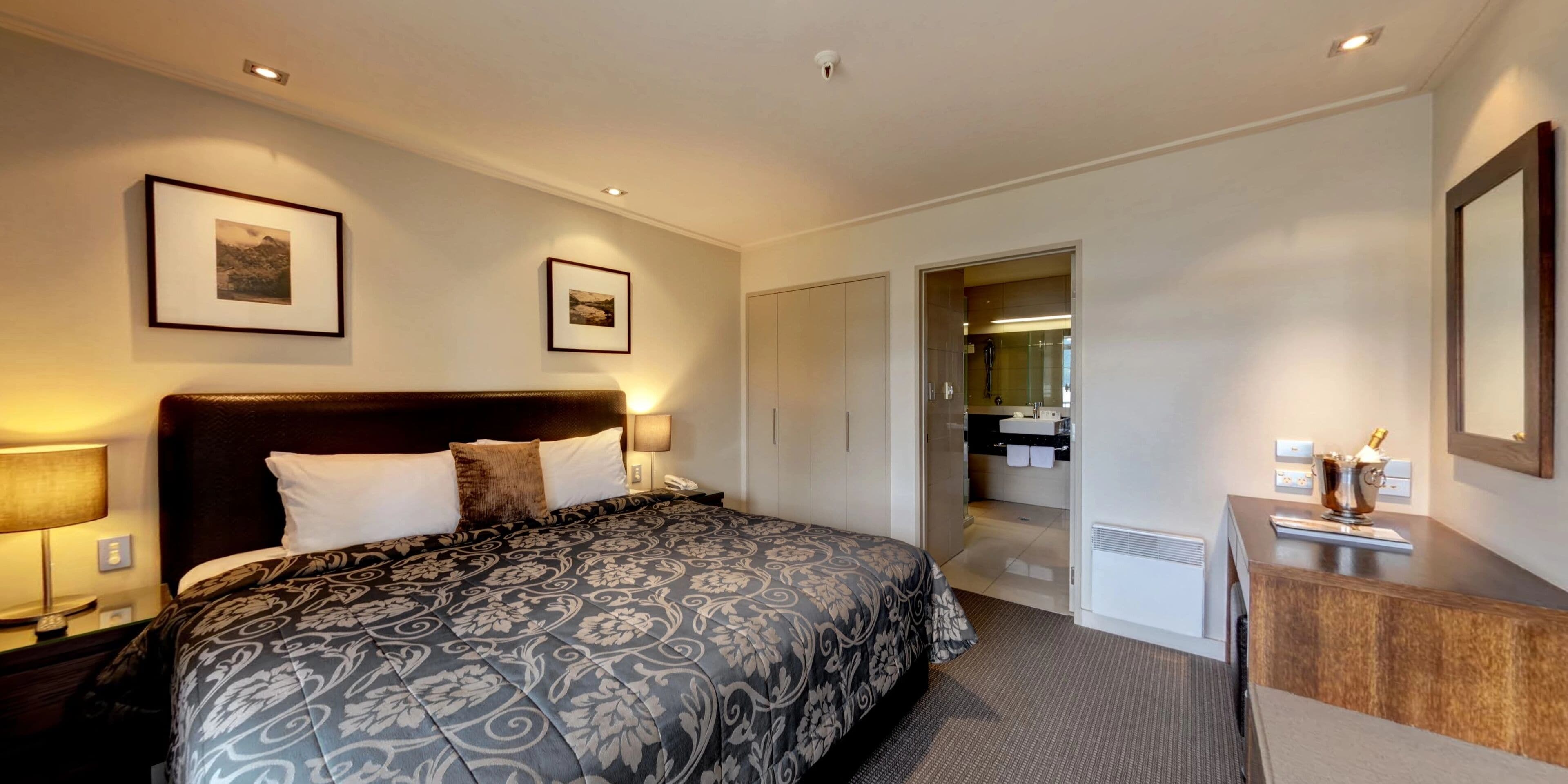 A hotel room with a large bed, two framed pictures on the wall, a desk with a minibar, and a bathroom visible through an open door.