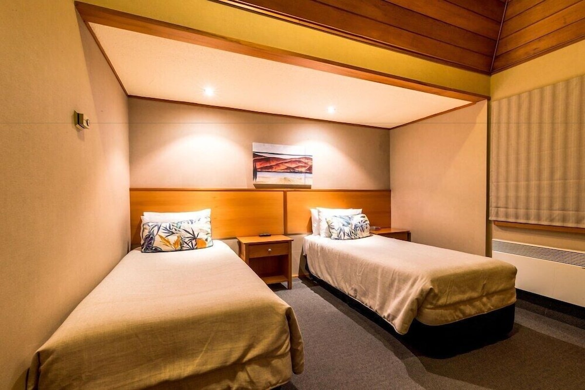A hotel room with two beds, a nightstand, a painting on the wall, and a window with blinds.
