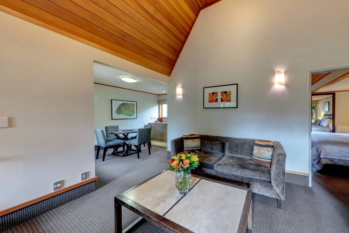 A spacious living area with a wooden ceiling, a sofa, a coffee table with flowers, a dining area with chairs, and a kitchen in the background.