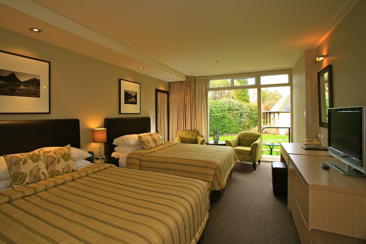 A hotel room with two beds, a desk with a TV, a chair, and a window with a view of greenery.