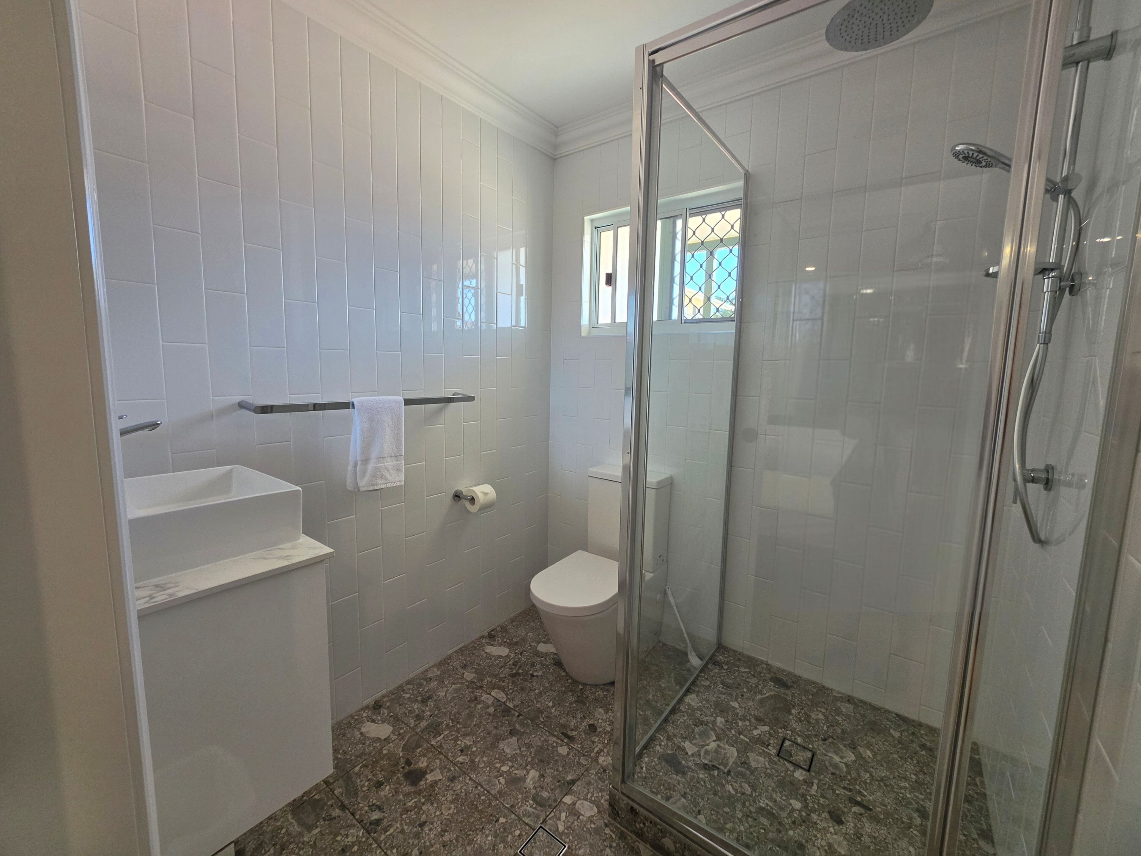 A bathroom with white tiled walls, a glass-enclosed shower, a toilet, and a sink with a towel hanging on the wall.