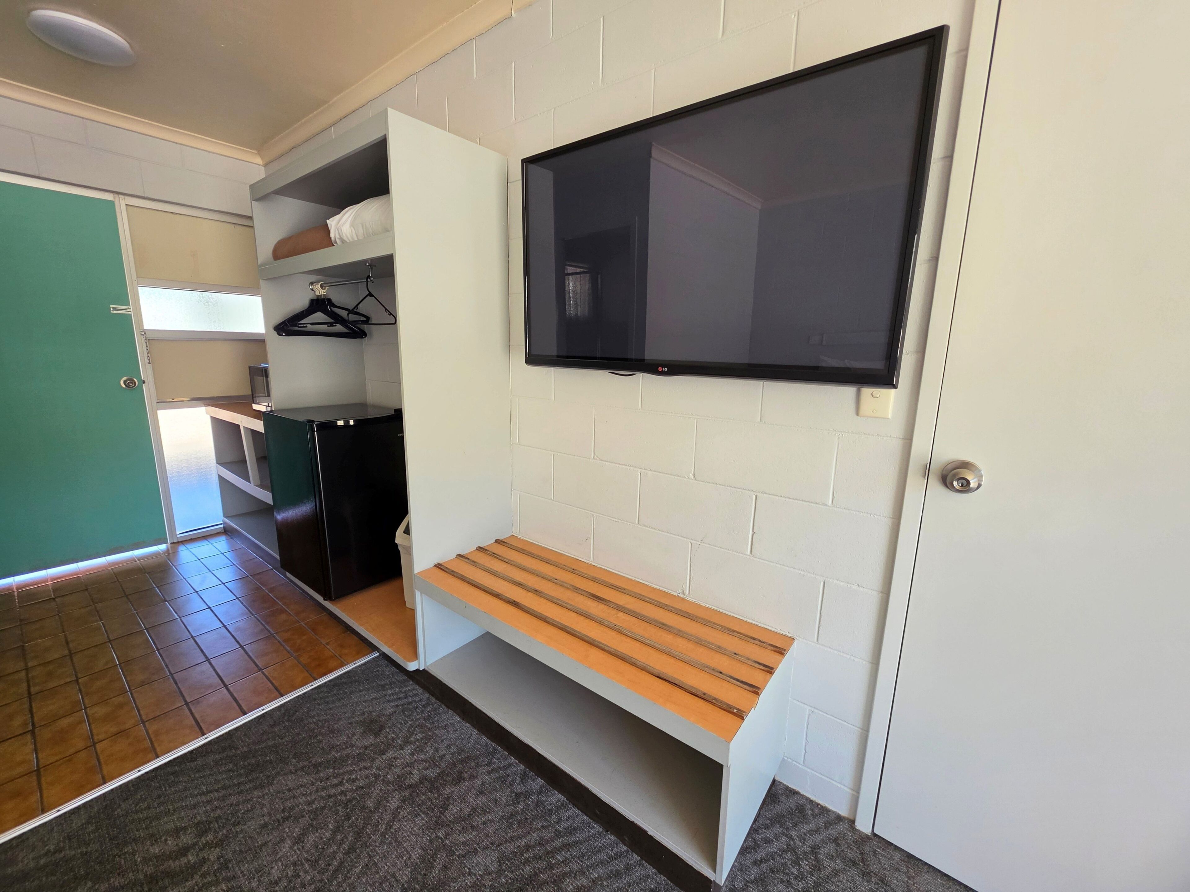 A room with a flat-screen TV mounted on the wall, a bench, a small refrigerator, and a door leading to another room.