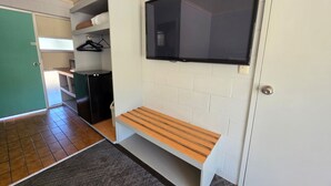 Habitación con una televisión de pantalla plana montada en la pared, un banco, un refrigerador pequeño y una puerta que da a otra habitación.