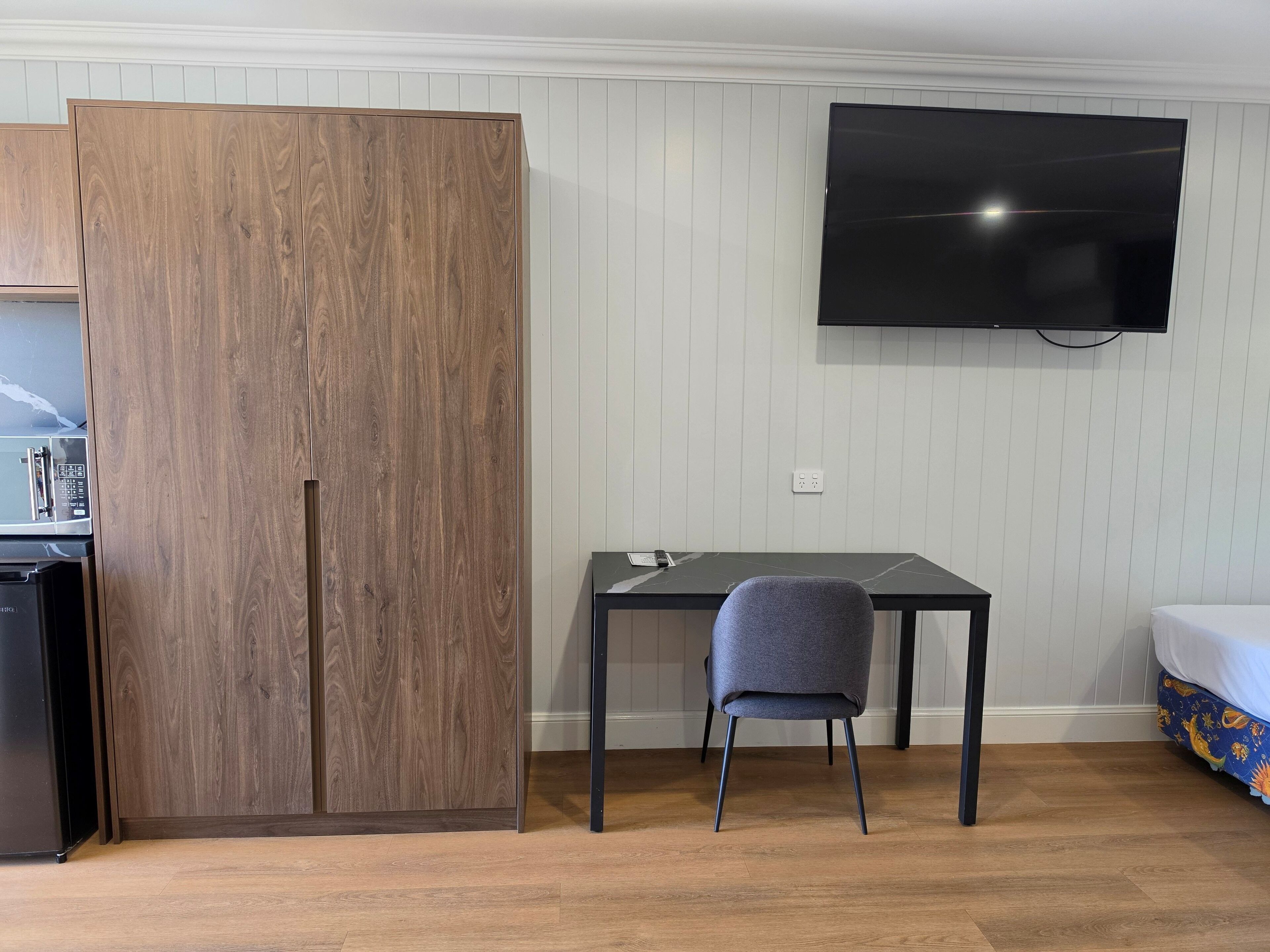 A room with a wooden wardrobe, a desk with a chair, a bed with a patterned cover, and a mounted TV.