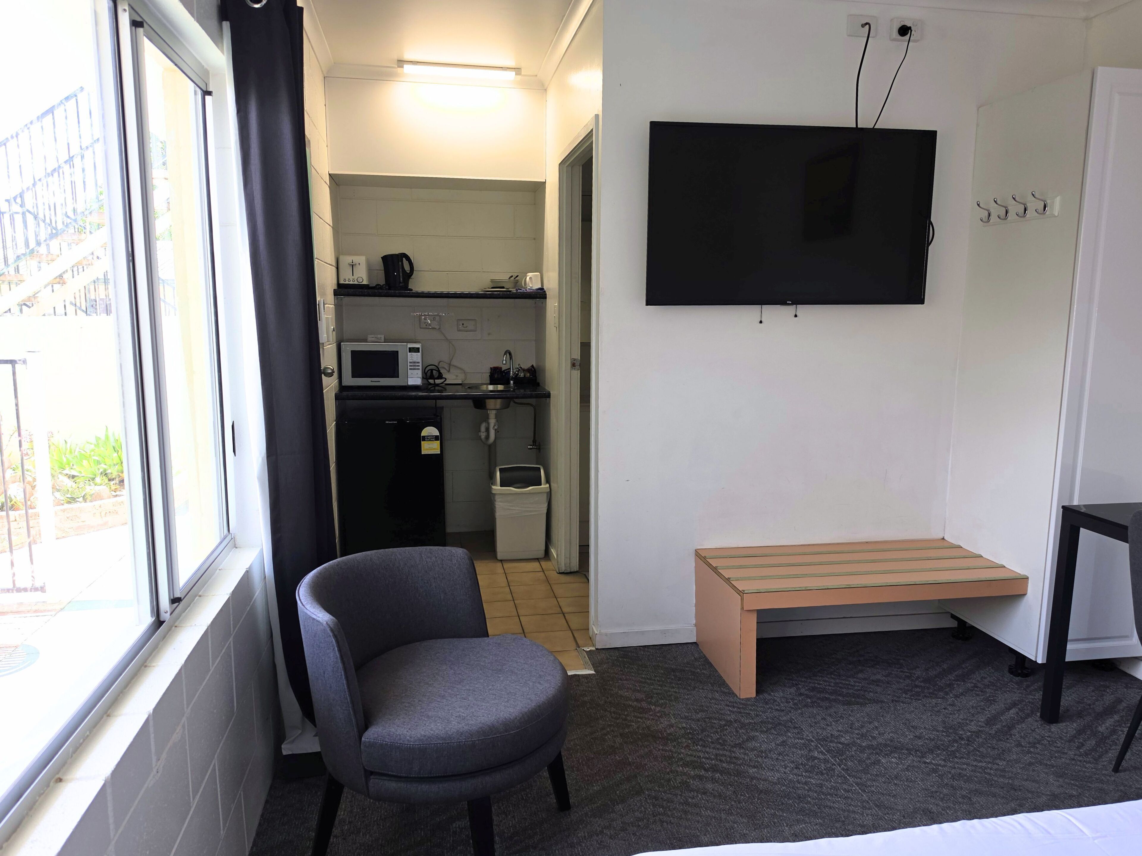 A compact room with a single grey chair, a small wooden bench, a television mounted on the wall, and a kitchenette in the background.