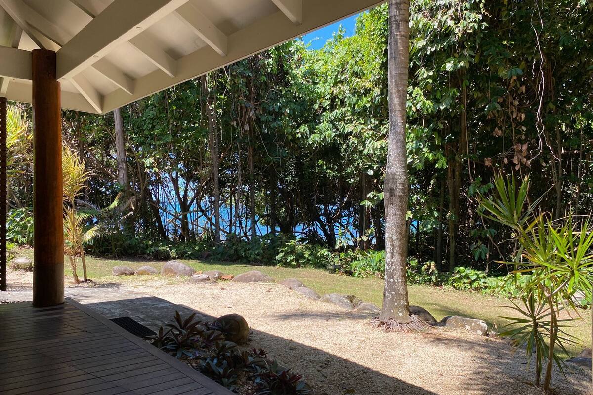 A covered outdoor area with a wooden deck, surrounded by dense greenery and a view of a body of water.