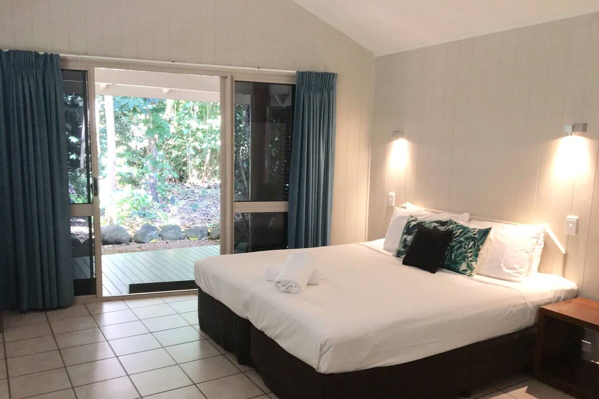 A hotel room with a large bed, a sliding glass door leading to a balcony, and a nightstand with a lamp.
