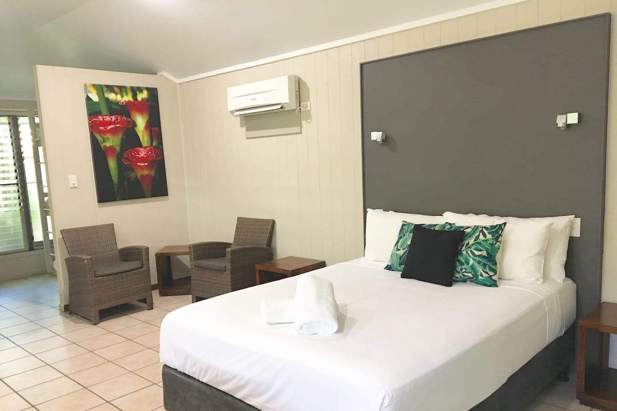 A hotel room with a large bed, two chairs, a small table, a painting, and an air conditioning unit.
