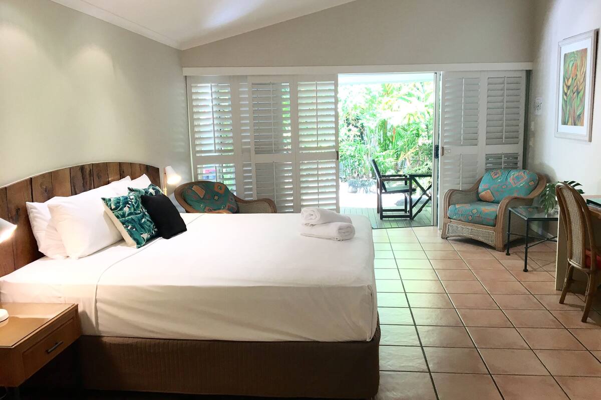 A hotel room with a large bed, a wooden headboard, a nightstand, a chair with a patterned cushion, and a view of the outdoors through a door with shutters.