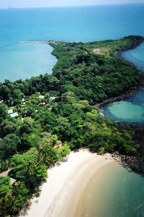 An aerial view of a coastal area with a sandy beach, dense greenery, and a clear blue sea.