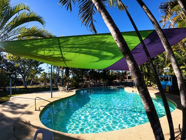 Ein Pool mit geschwungener Kante, umgeben von Palmen und beschattet durch eine grüne und violette Markise.