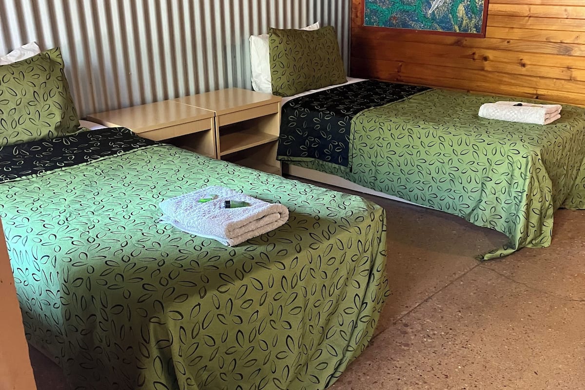 Ein Zimmer mit zwei Betten, jeweils mit grün gemusterten Bettbezügen, einem Holzkopfstück, einem kleinen Tisch und einem Gemälde an der Wand.
