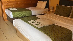 A room with two single beds, each with a green patterned bedspread and white pillows. There is a nightstand with a lamp and a small table with a lamp. The room has wooden walls and a tiled floor. - Territory Manor Motel & Caravan Park (Mataranka)