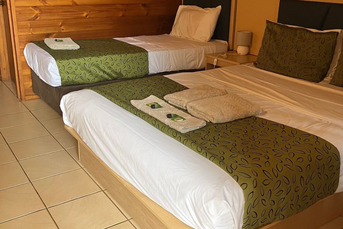 Ein Zimmer mit zwei Einzelbetten, jeweils mit einer grün gemusterten Tagesdecke und weißen Kissen. Ein Nachttisch mit Lampe sowie ein kleiner Tisch mit Lampe sind vorhanden. Die Wände sind aus Holz und der Boden ist mit Fliesen belegt.