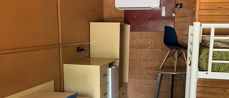 Una habitación compacta con una cama litera, un escritorio pequeño con una silla, una mesa con sillas y un aire acondicionado instalado en la pared.