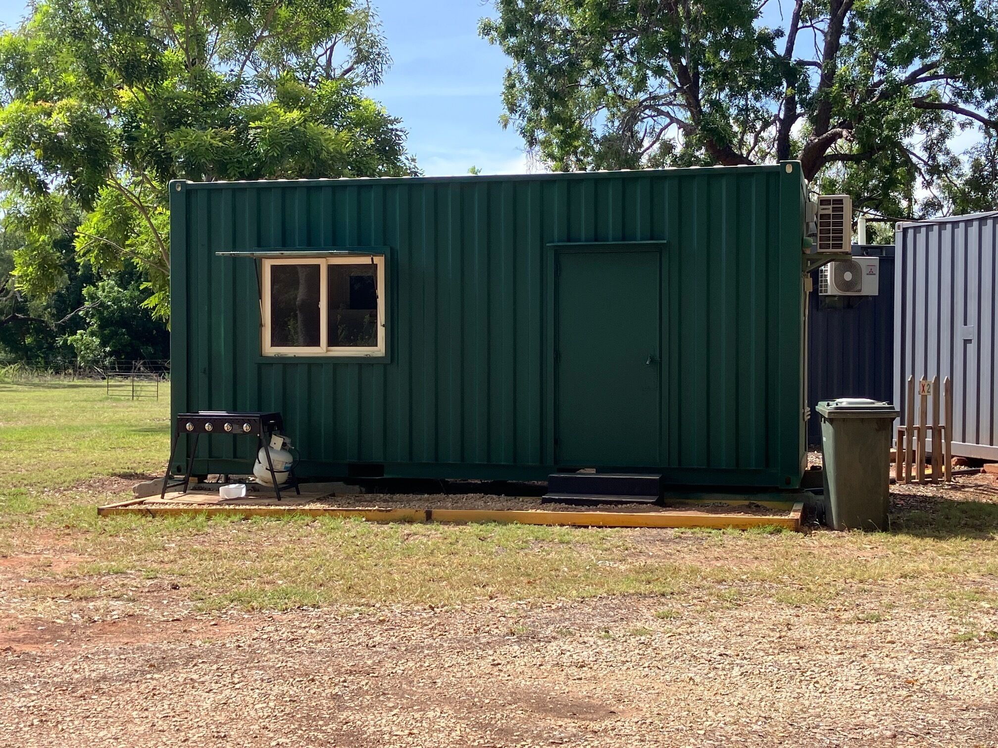 A small, green, single-story building with a window and a door, situated in a grassy area with trees in the background.