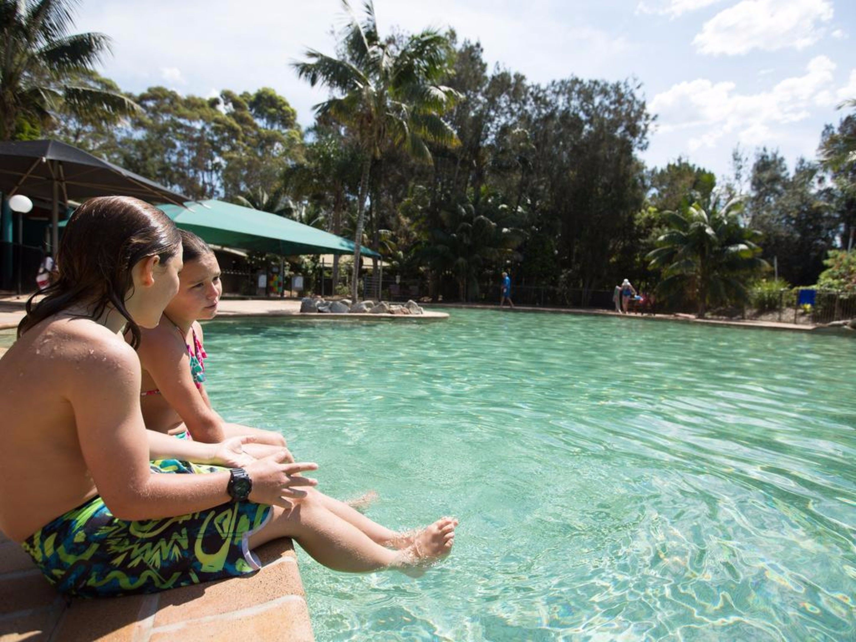 Zona de piscina con agua cristalina, rodeada de palmeras y un área de descanso cubierta.