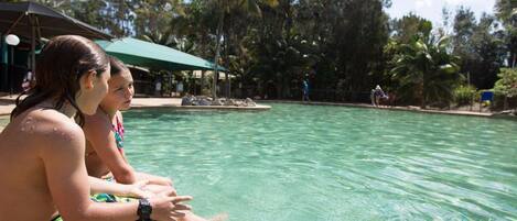 Zona de piscina con agua cristalina, rodeada de palmeras y un área de descanso cubierta.