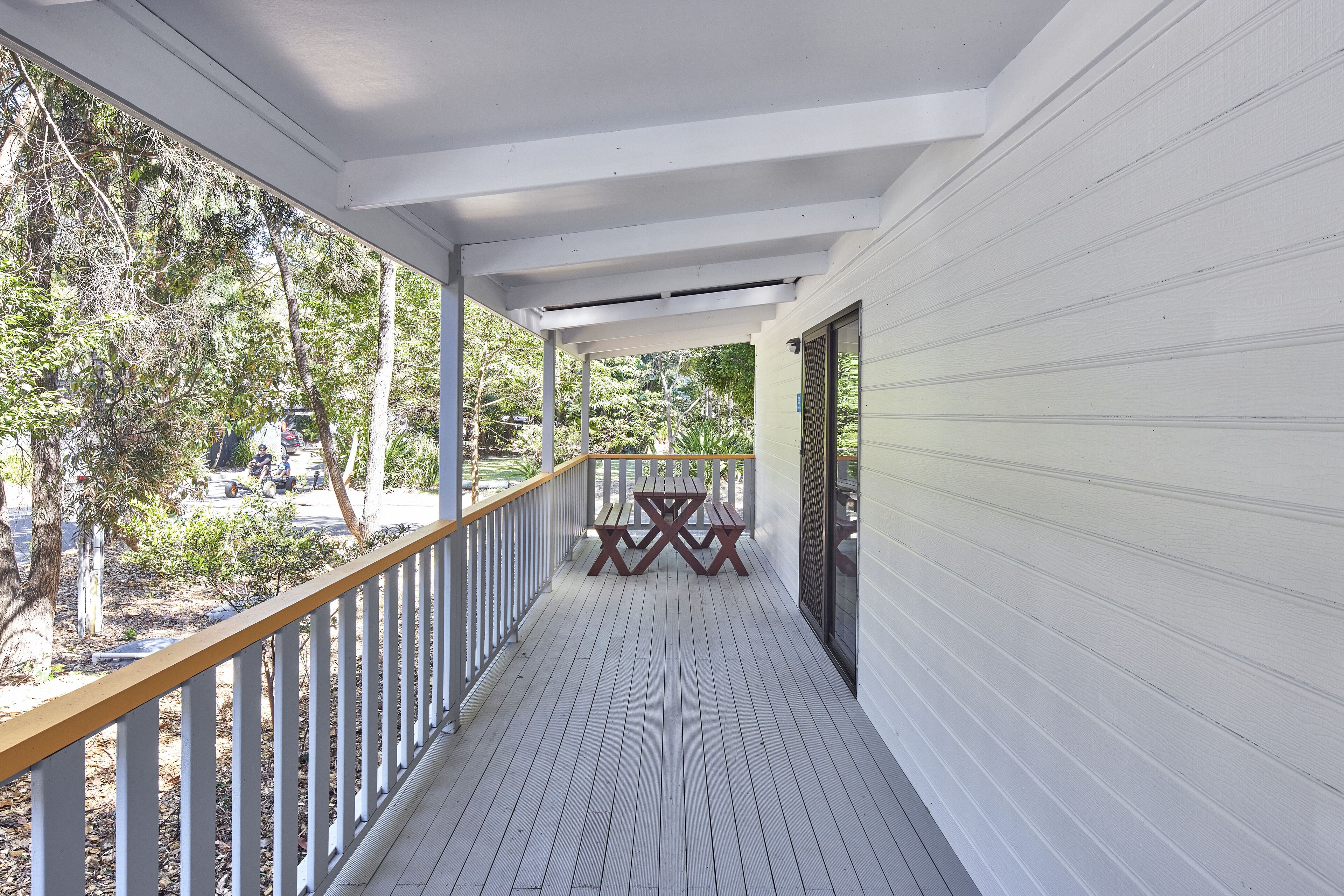 A covered porch with a wooden railing, a picnic table, and a view of a garden with trees and a lawn.