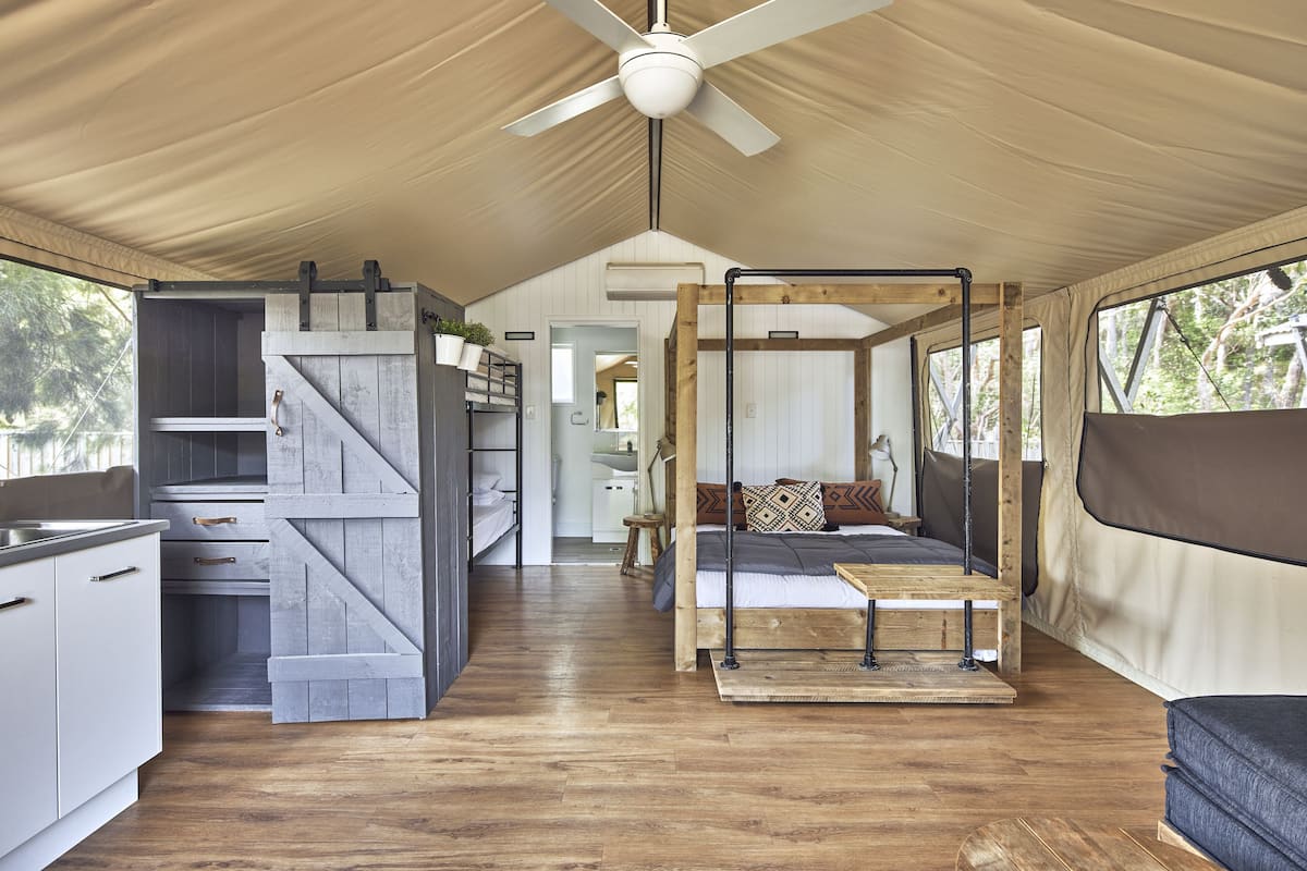 A tented accommodation with a wooden interior, a bed with a headboard, a dining area, and a kitchenette.