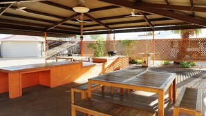 Un patio al aire libre con mesas y bancos de madera, una zona cubierta con ventilador de techo y vista a una calle en el fondo.