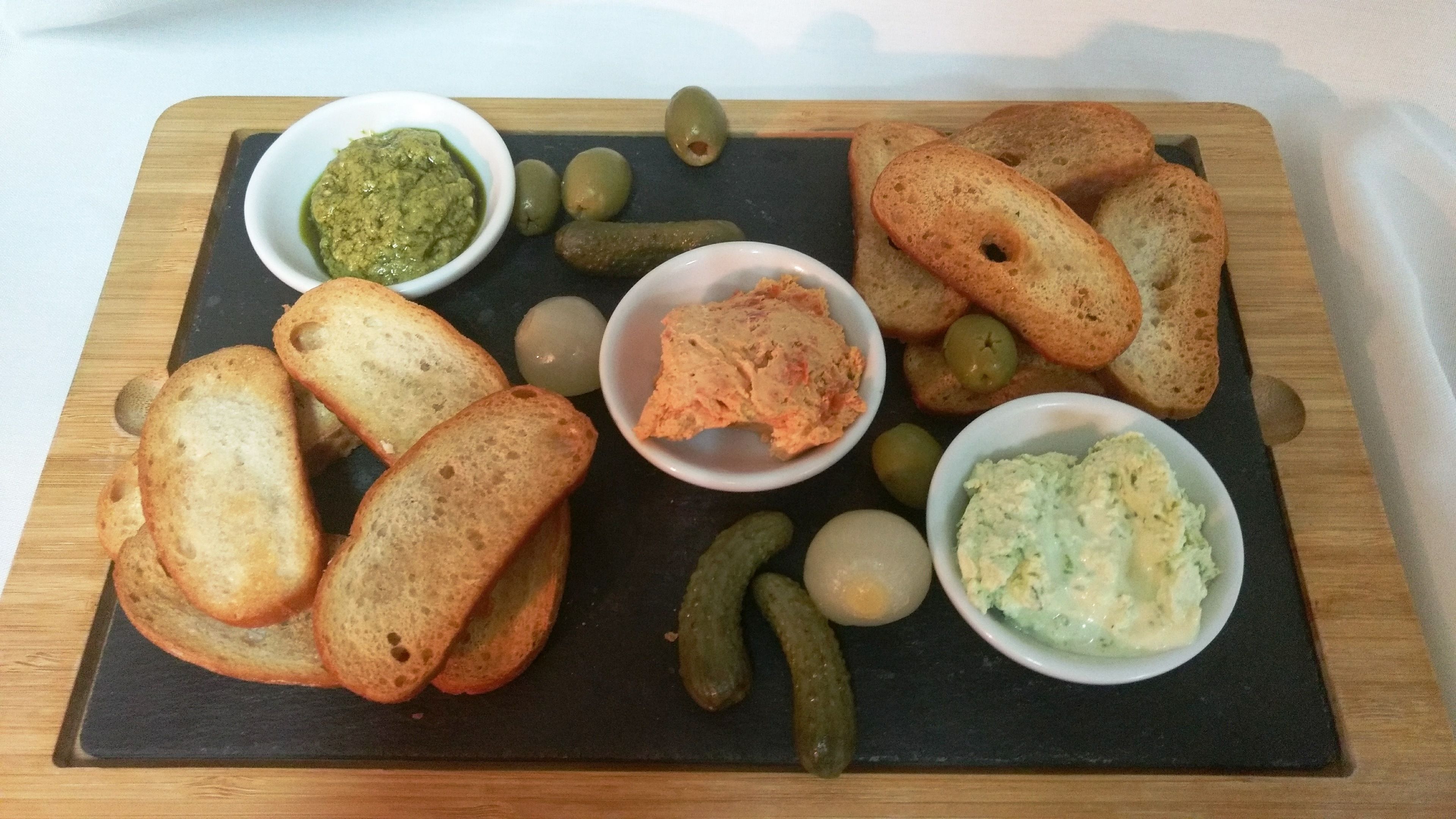 A wooden board with various food items including bread, olives, pickles, and dips.