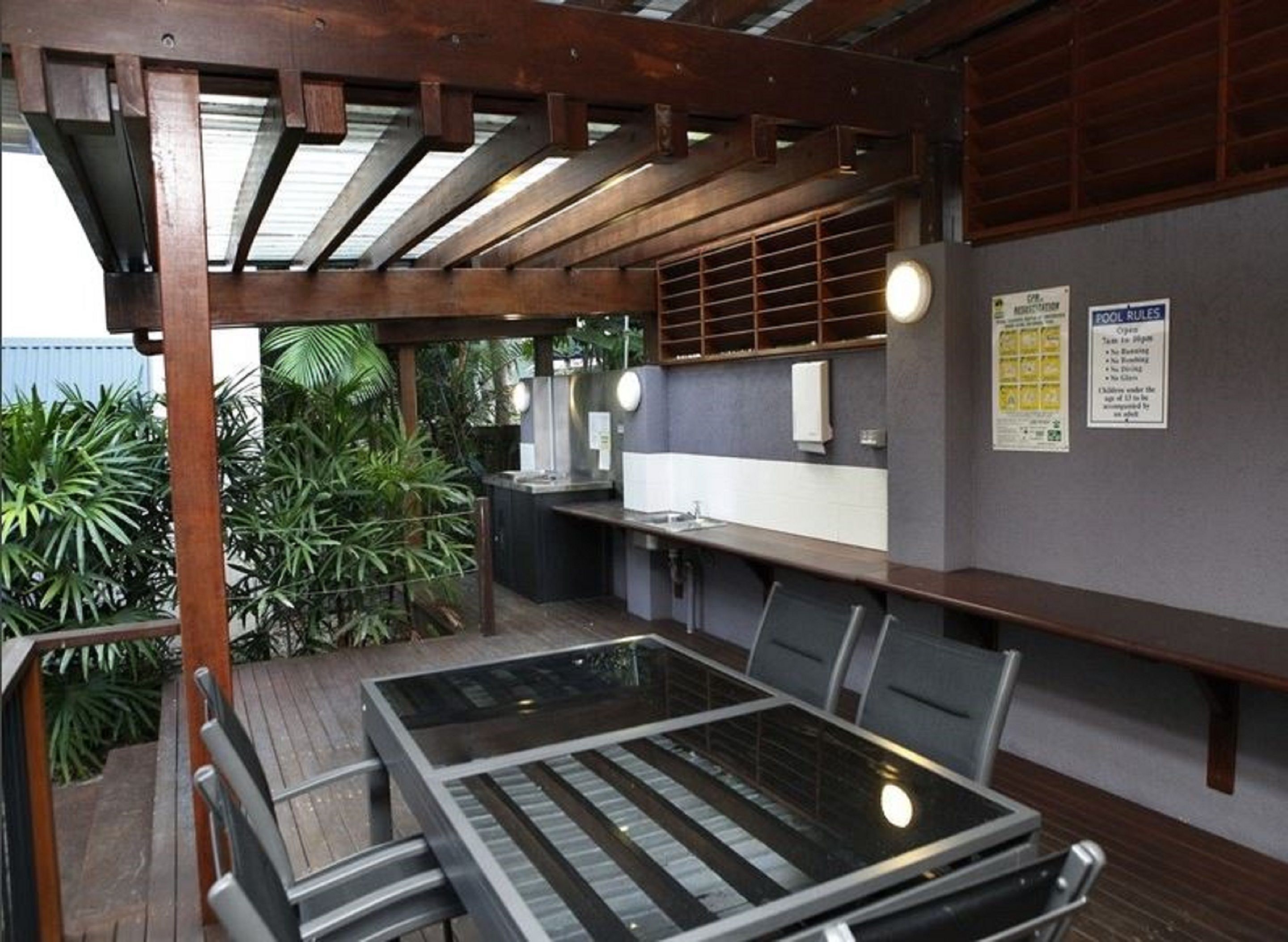 Een terras buiten met een houten pergola, een tafel met glazen blad en stoelen. Er is een geïntegreerde keuken met een spoelbak en een bord met regels voor het zwembad.