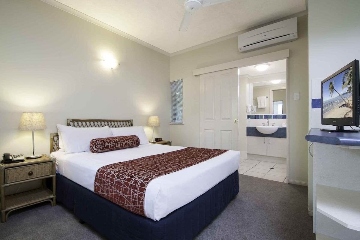 A hotel room with a bed, bedside lamps, a TV, a bathroom with a sink, and an air conditioning unit.