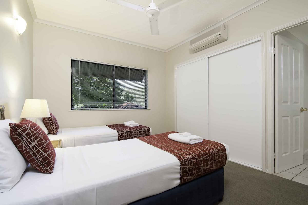A hotel room with two beds, a window with blinds, a ceiling fan, an air conditioning unit, and a wardrobe.
