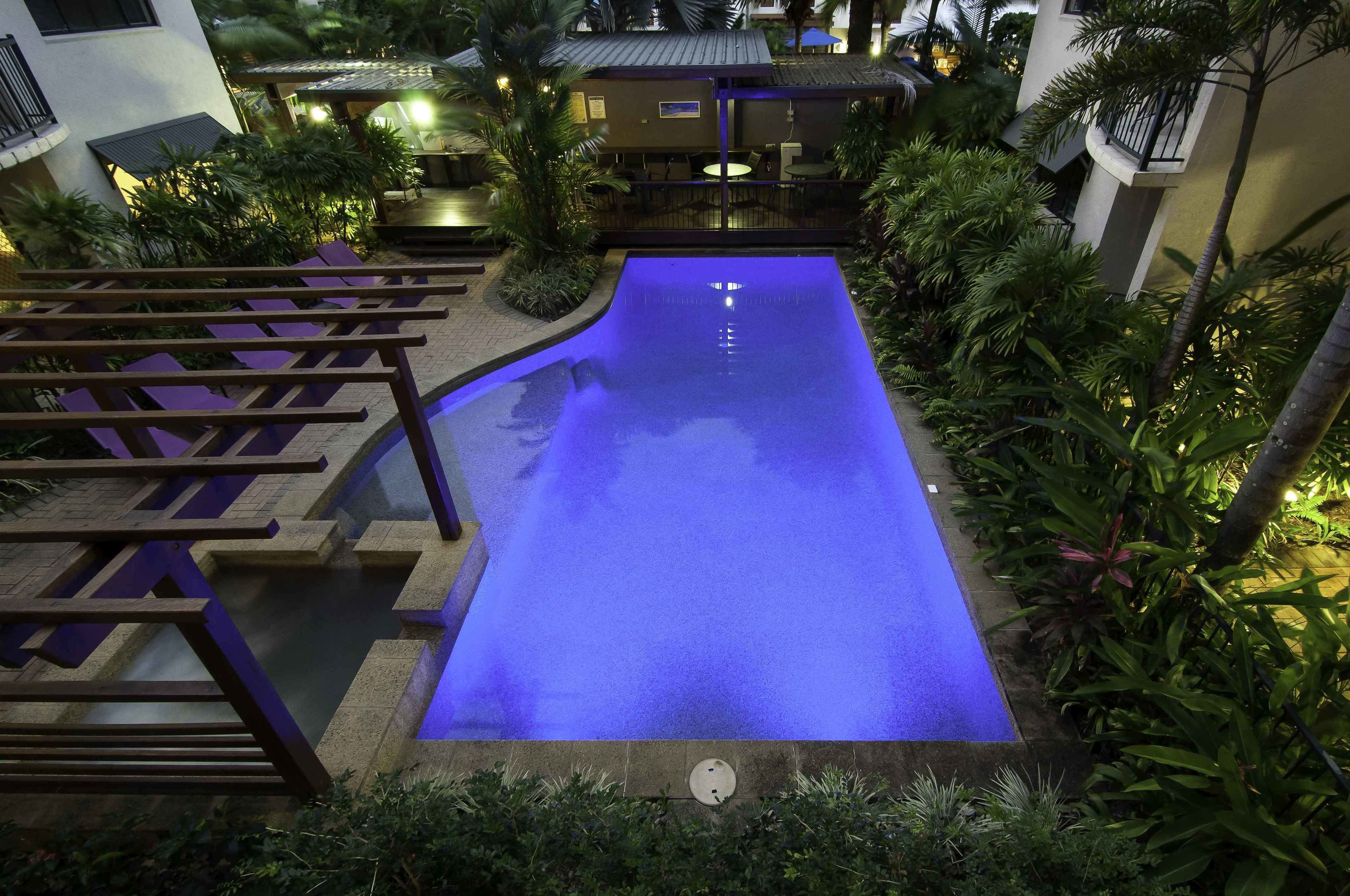 Una piscina con un deck iluminado en azul, rodeada de vegetación exuberante y una pequeña zona de estar al aire libre.