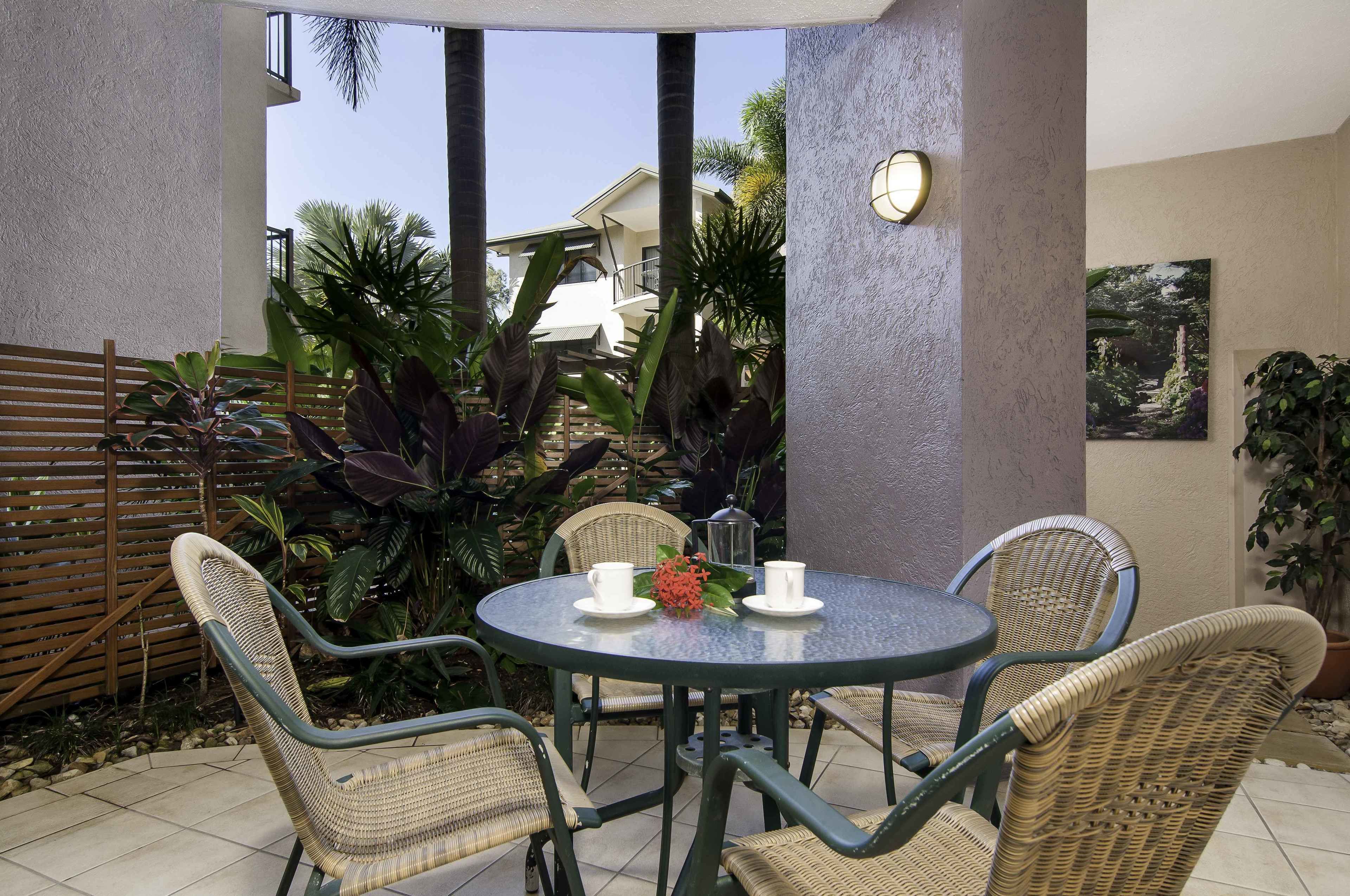 Un patio con sillas de mimbre y una mesa, rodeado de plantas y con vistas a un edificio.