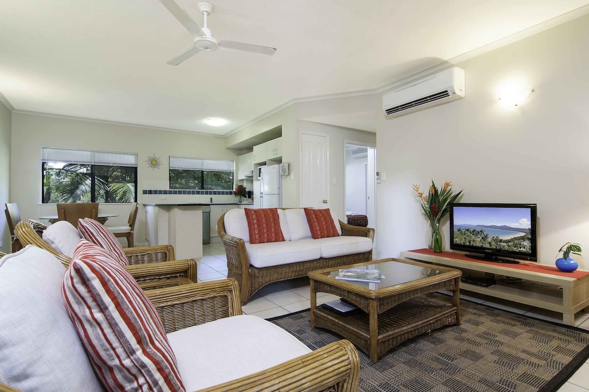A living room with wicker furniture, a television, and a kitchen in the background.