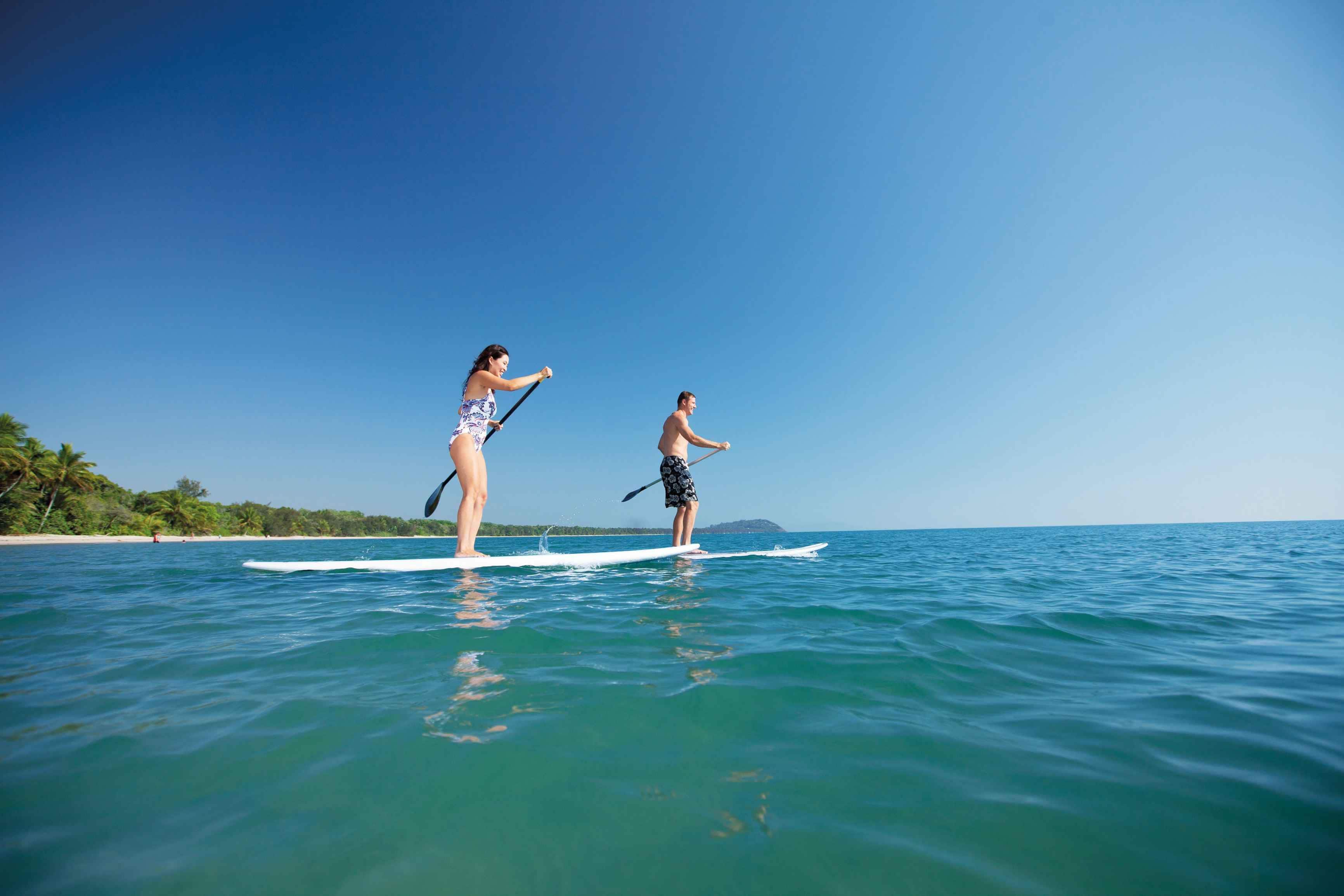 Two people paddleboarding on a calm sea with a clear sky and a distant shoreline with vegetation.