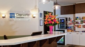 A reception area with a sign for the Clearwater Suite Hotel, a white reception desk, a pink vase with flowers, and a kitchen area with cabinets and a refrigerator. - Clearwater Suite Hotel (Fort McMurray)