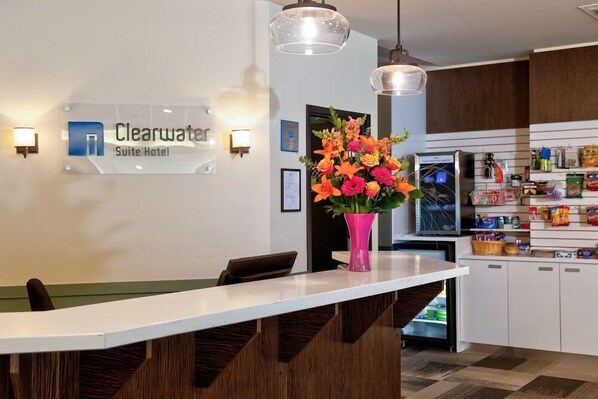 A reception area with a sign for the Clearwater Suite Hotel, a white reception desk, a pink vase with flowers, and a kitchen area with cabinets and a refrigerator.