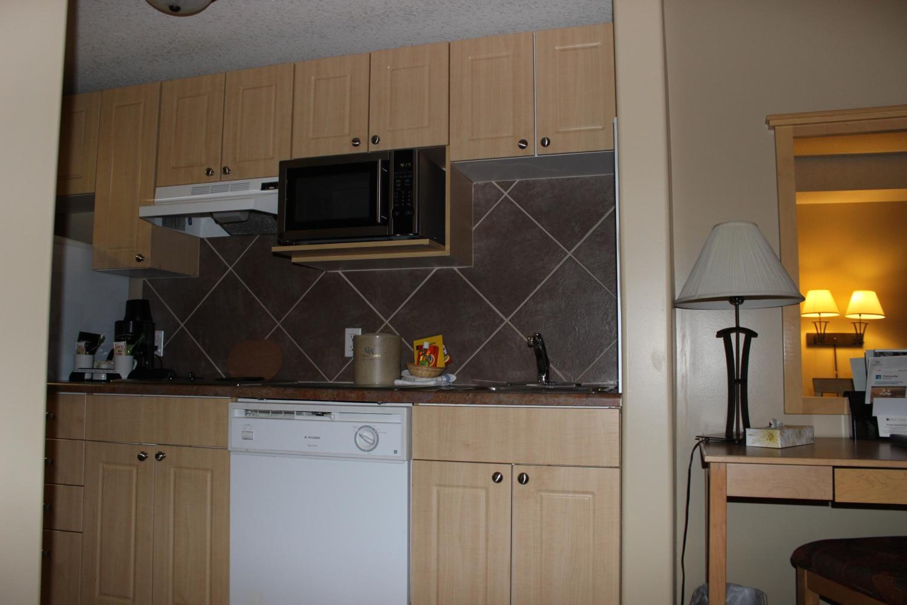 A compact kitchen with wooden cabinets, a microwave, a dishwasher, and a small table with a lamp.