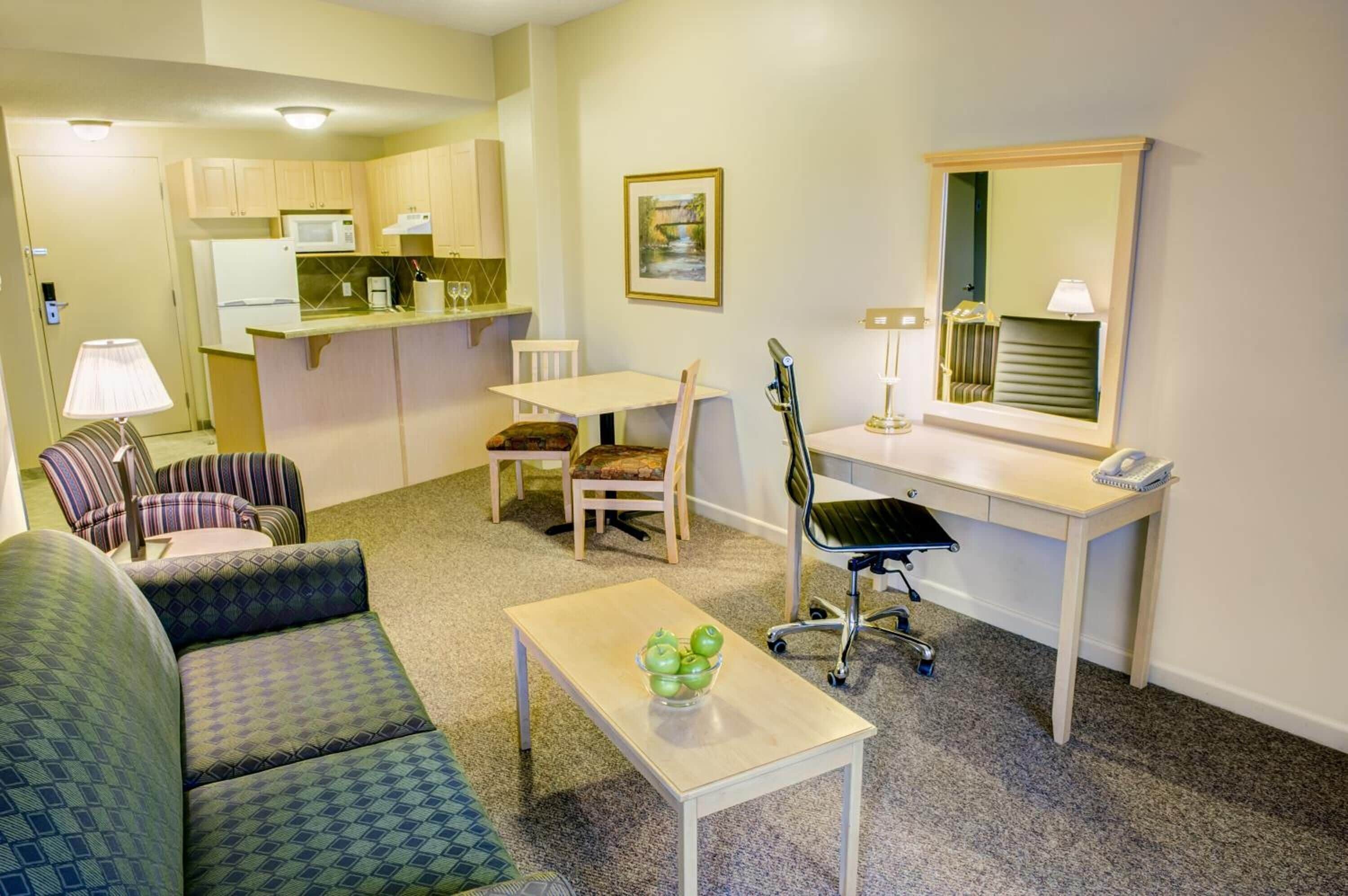 A compact living space with a kitchenette, a dining area, a seating area with a sofa and chairs, a desk with a chair, and a small table with a lamp.