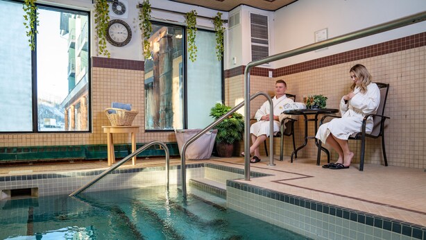 An indoor pool area with a clear view of the outside, a seating area with two people, and a small table with drinks. - Clearwater Suite Hotel (Fort McMurray)