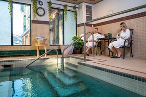 An indoor pool area with a clear view of the outside, a seating area with two people, and a small table with drinks. - Clearwater Suite Hotel (Fort McMurray)