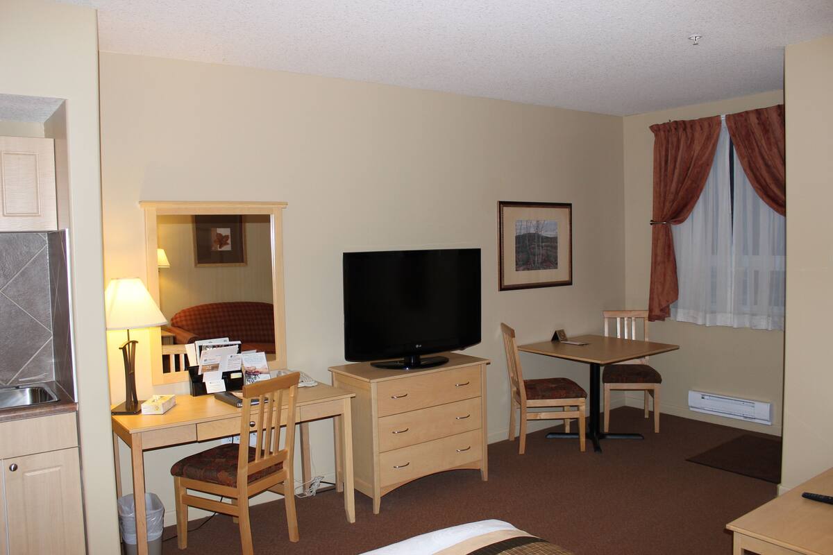 A hotel room with a kitchenette, a flat-screen TV, a desk with a chair, a dining table with chairs, and a bed.