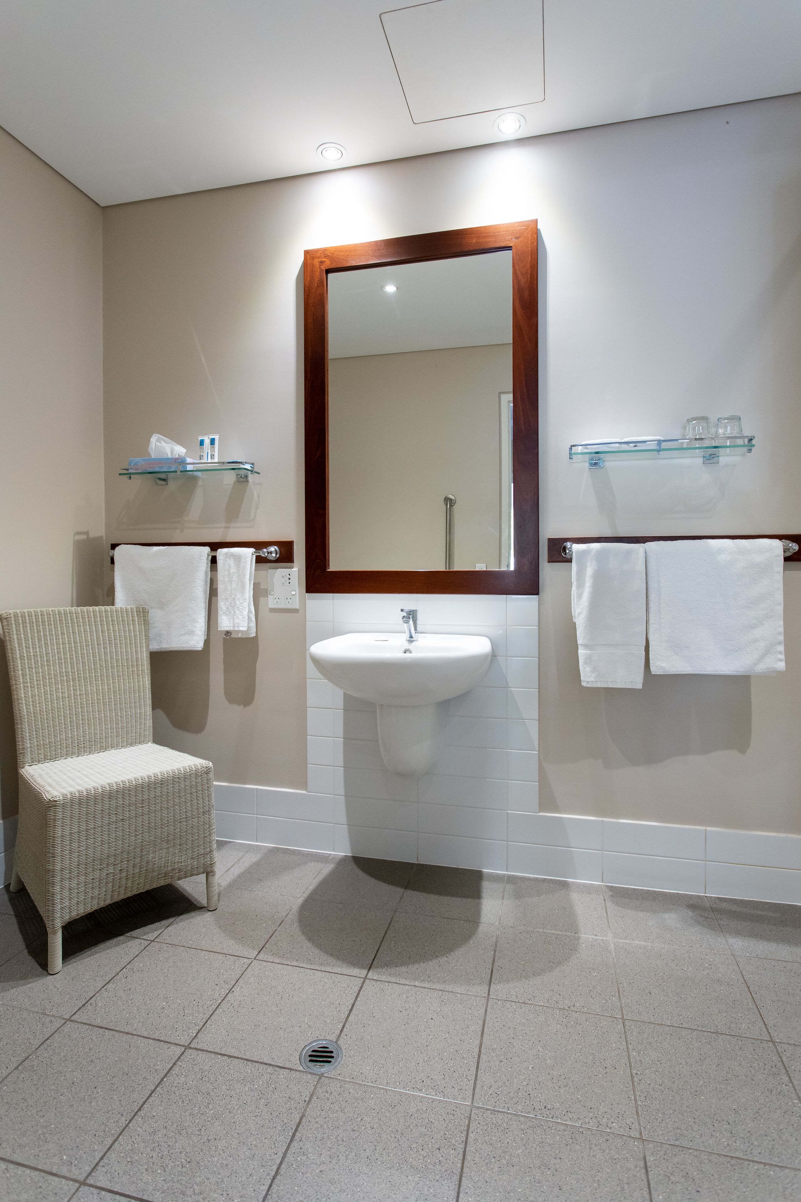 Une salle de bain avec un lavabo blanc, un miroir avec un cadre en bois, deux porte-serviettes, une chaise et une étagère en verre contenant des articles de toilette.