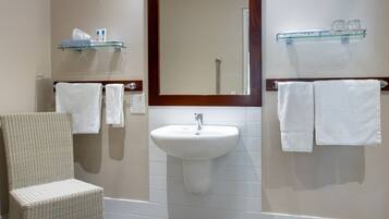 Une salle de bain avec un lavabo blanc, un miroir avec un cadre en bois, deux porte-serviettes, une chaise et une étagère en verre contenant des articles de toilette.