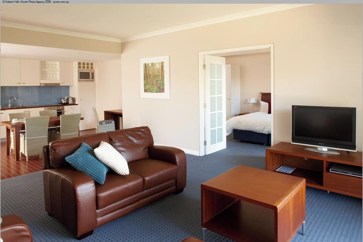 Una habitación de hotel moderna con un sofá de cuero marrón, un televisor de pantalla plana, una mesa de centro de madera y una zona de comedor con una cocina al fondo.