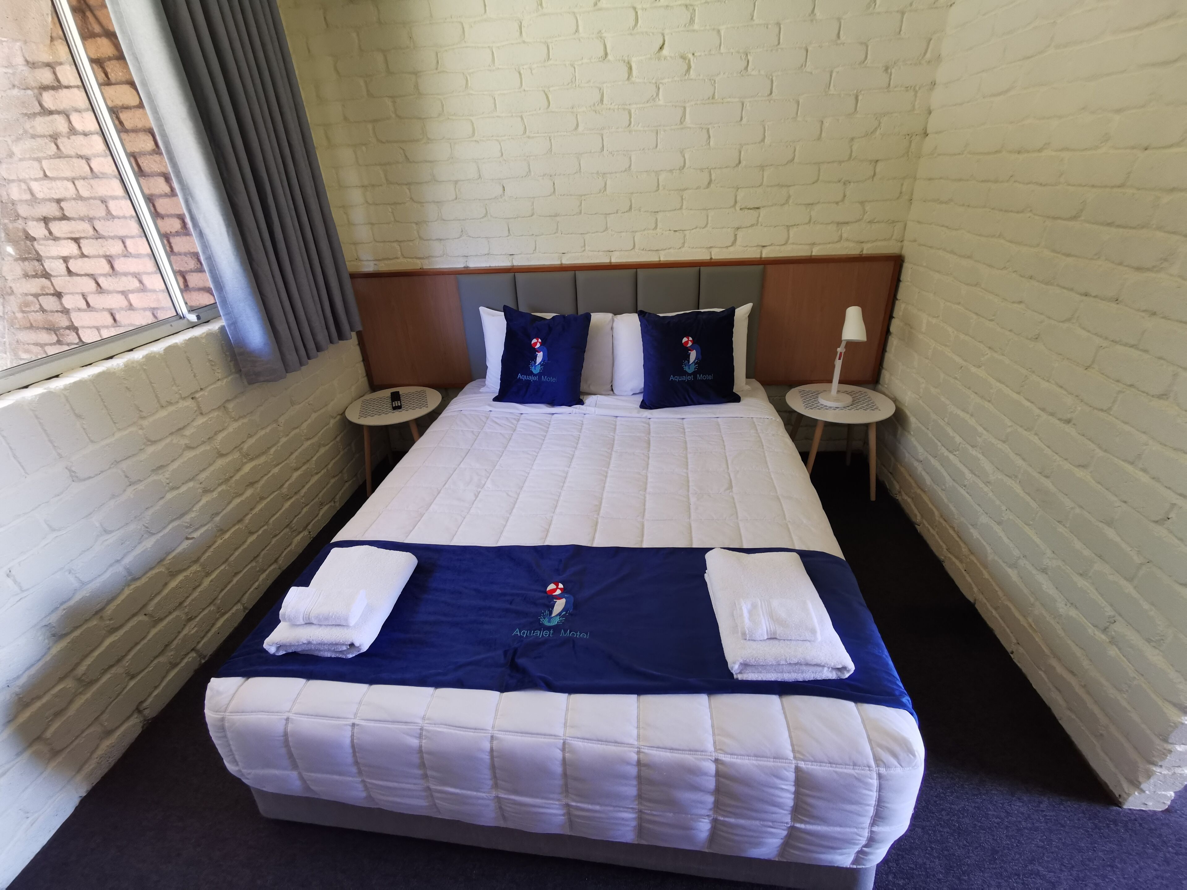 A hotel room with a brick wall, a large bed with white and blue bedding, two pillows with a logo, a small round table, and a lamp.